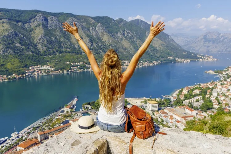 Eine Frau sitzt mit ausgebreiteten Armen auf einer Mauer und blickt auf eine tiefblaue Bucht, umgeben von Bergen und einer Küstenstadt mit roten Dächern.