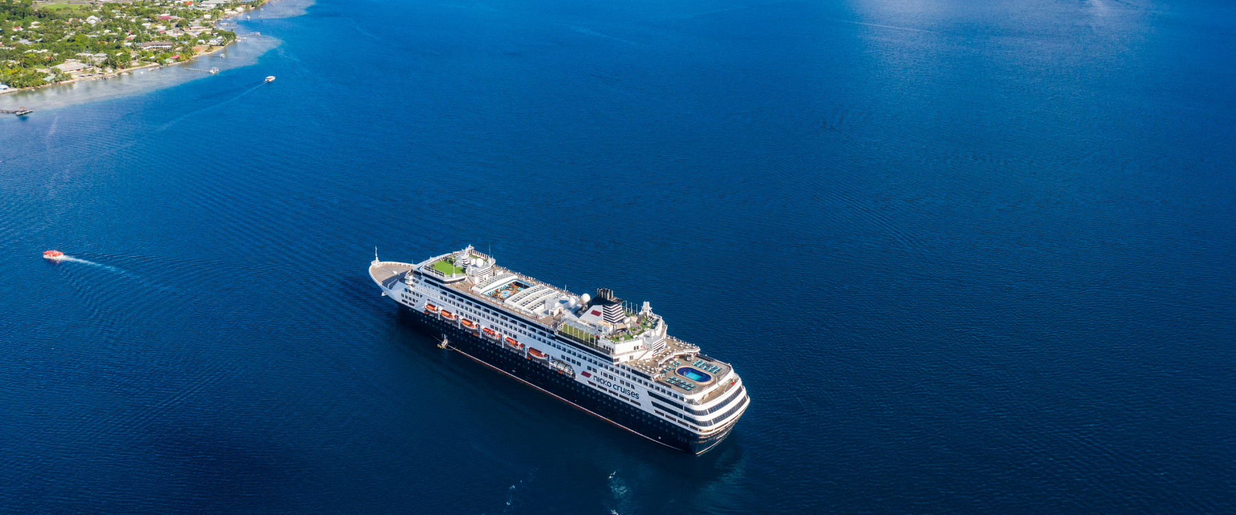 Ein großes Kreuzfahrtschiff der Reederei nicko Cruises fährt über tiefblaues, ruhiges Meer. Auf dem Schiff sind mehrere Decks mit Pools, Liegeflächen und Freizeitbereichen sichtbar. Am oberen linken Rand ist die Küste mit Häusern und grüner Vegetation zu sehen, außerdem fahren zwei kleinere Boote auf dem Wasser.